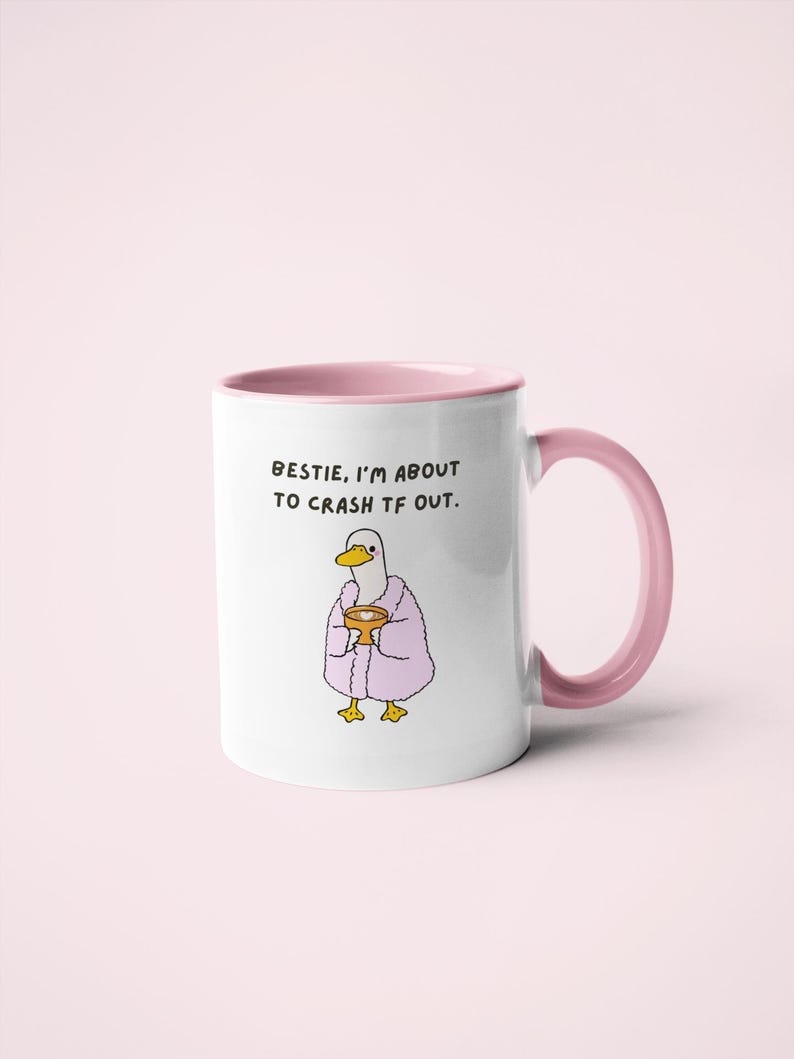 May include: White ceramic mug with a pink interior and handle. The mug features a cartoon duck wearing a pink robe and holding a golden cup. Text on the mug reads, "BESTIE, I'M ABOUT TO CRASH TF OUT."