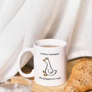 Puede incluir: Taza de cer&aacute;mica blanca con una ilustraci&oacute;n de pato de dibujos animados y el texto "I'M BUILT DIFFERENT LIKE INCORRECTLY I THINK". La taza est&aacute; sobre una tabla de madera con un cruas&aacute;n y un vaso de agua.