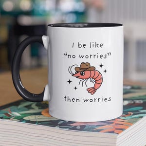 May include: White ceramic mug with a black handle and rim. The mug features a cartoon shrimp wearing a cowboy hat, with the text "I be like 'no worries' then worries".