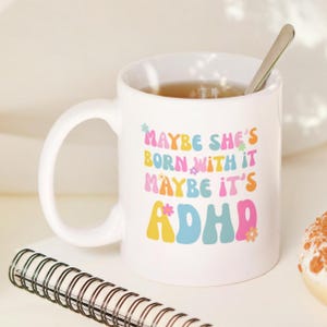 May include: White ceramic mug with the text "Maybe she's born with it, maybe it's ADHD" in colorful, retro-style lettering. A silver spoon rests inside the mug, which is filled with a light brown liquid. A spiral notebook is in the foreground.