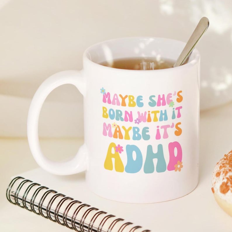 May include: White ceramic mug with the text "Maybe she's born with it, maybe it's ADHD" in colorful, retro-style lettering. A silver spoon rests inside the mug, which is filled with a light brown liquid. A spiral notebook is in the foreground.