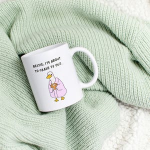 May include: White ceramic mug with a humorous illustration of a duck in a pink robe holding a bowl. The mug has the text "BESTIE, I'M ABOUT TO CRASH TF OUT." The mug is resting on a light green knitted blanket.