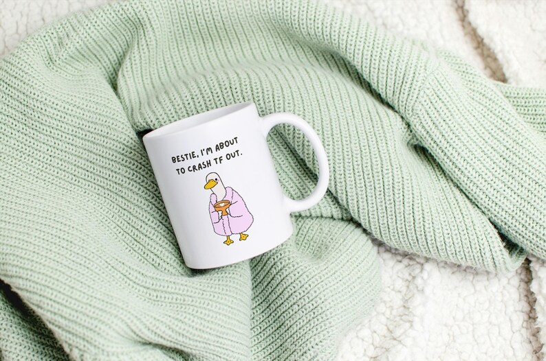May include: White ceramic mug with a humorous illustration of a duck in a pink robe holding a bowl. The mug has the text "BESTIE, I'M ABOUT TO CRASH TF OUT." The mug is resting on a light green knitted blanket.