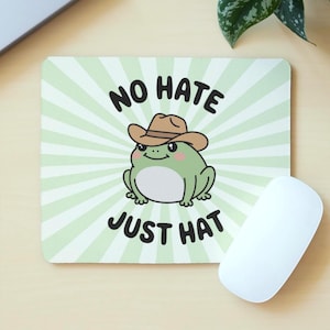 May include: A rectangular mouse mat featuring a cartoon frog in a cowboy hat. The frog is green with pink cheeks, set against a light green and white striped background. Text reads "NO HATE JUST HAT". A white computer mouse is on the right.