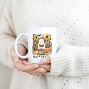 May include: White ceramic mug featuring a whimsical illustration of a ghost reading a book, surrounded by sunflowers, pumpkins, and bats. The text "READ MORE" is printed below the image. A perfect gift for book lovers.