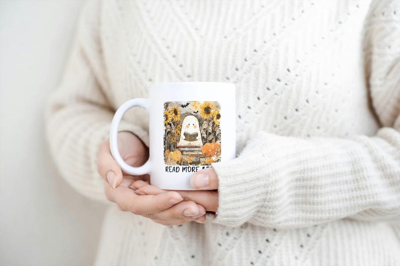 May include: White ceramic mug featuring a whimsical illustration of a ghost reading a book, surrounded by sunflowers, pumpkins, and bats. The text "READ MORE" is printed below the image. A perfect gift for book lovers.