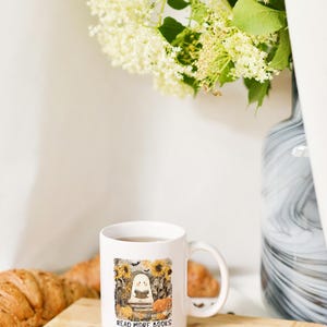 May include: White ceramic mug with a handle, featuring a fall-themed illustration of a ghost reading a book, surrounded by pumpkins and sunflowers. The text "READ MORE BOOKS" is below the illustration. The mug sits on a wooden board.