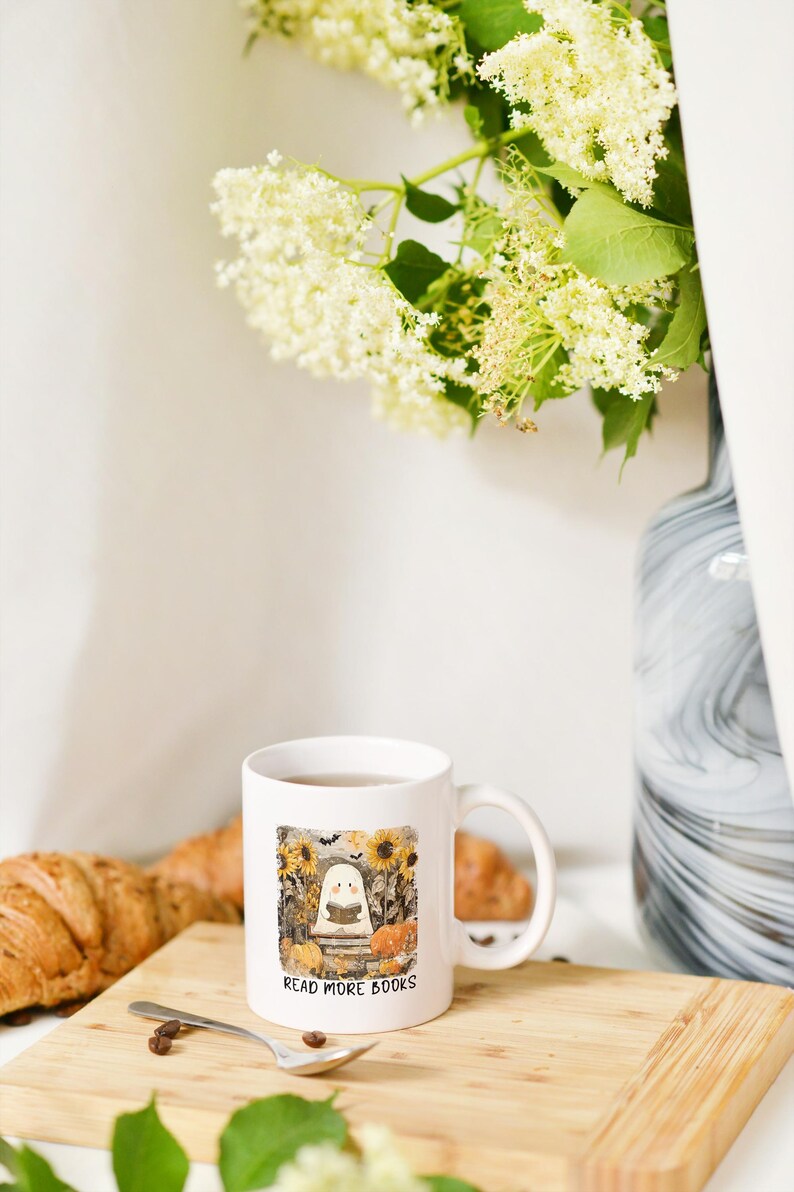 May include: White ceramic mug with a handle, featuring a fall-themed illustration of a ghost reading a book, surrounded by pumpkins and sunflowers. The text "READ MORE BOOKS" is below the illustration. The mug sits on a wooden board.