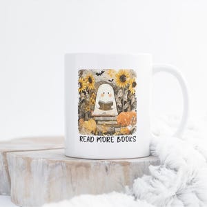 May include: White ceramic mug featuring a ghost reading a book, surrounded by sunflowers and pumpkins. The mug has the text "READ MORE BOOKS" printed on it. The design has a fall or Halloween theme.