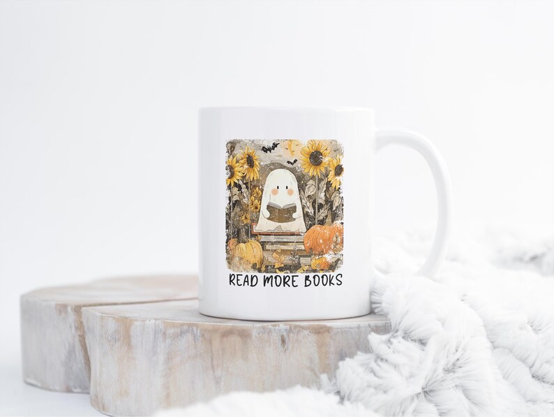 May include: White ceramic mug featuring a ghost reading a book, surrounded by sunflowers and pumpkins. The mug has the text "READ MORE BOOKS" printed on it. The design has a fall or Halloween theme.