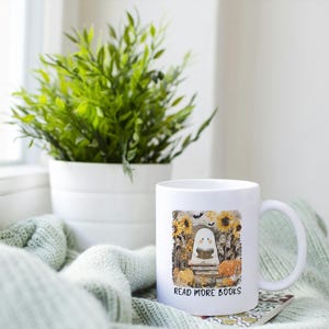 May include: White ceramic mug with a handle, featuring a colorful illustration of a ghost reading a book, surrounded by sunflowers and pumpkins. The text "READ MORE BOOKS" is printed below the image. A green plant in a white pot is in the background.