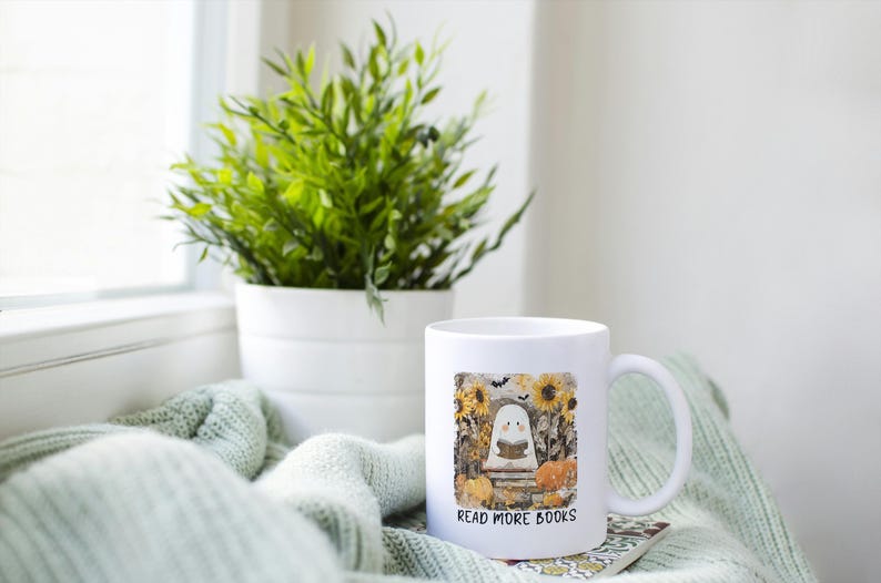 May include: White ceramic mug with a handle, featuring a colorful illustration of a ghost reading a book, surrounded by sunflowers and pumpkins. The text "READ MORE BOOKS" is printed below the image. A green plant in a white pot is in the background.