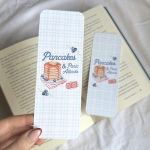 May include: Two rectangular bookmarks with a light blue plaid design. One is in focus, the other is blurred. The bookmarks display the text "Pancakes & Panic Attacks" with an illustration of pancakes, blueberries, and a red and white checkered napkin.