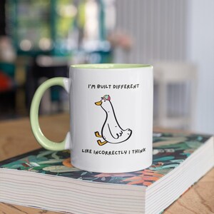 Puede incluir: Taza de cer&aacute;mica blanca con asa e interior verde claro. La taza presenta un pato de dibujos animados con sombrero y el texto "I'M BUILT DIFFERENT LIKE INCORRECTLY I THINK". La taza est&aacute; sobre un libro.