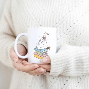 May include: White ceramic mug featuring a cartoon goose wearing a pink bow, perched atop a stack of colorful books. The mug has the text "SELF GOOSE BOOK CLUB" and a curved handle. A cozy, warm-toned sweater is in the background.
