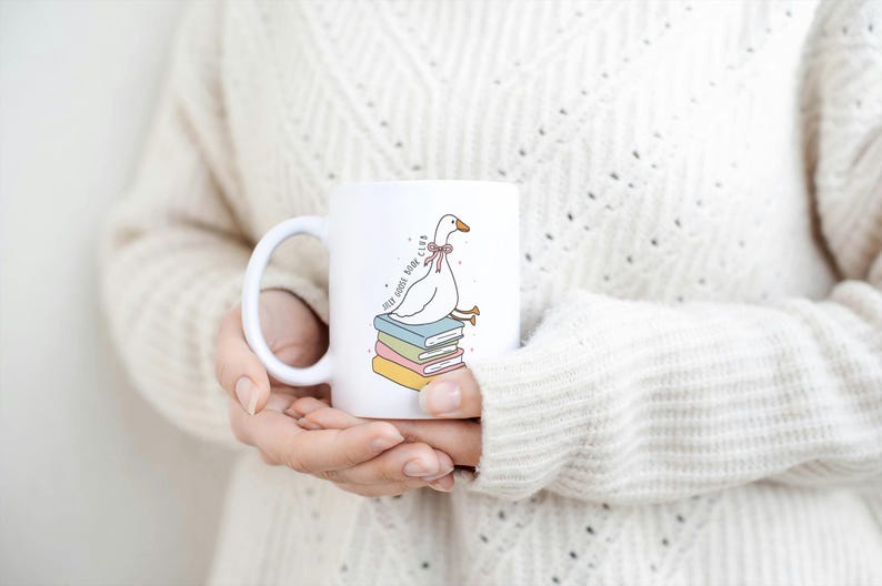 May include: White ceramic mug featuring a cartoon goose wearing a pink bow, perched atop a stack of colorful books. The mug has the text "SELF GOOSE BOOK CLUB" and a curved handle. A cozy, warm-toned sweater is in the background.