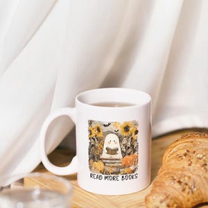 May include: White ceramic mug with a Halloween-themed illustration. The design features a cartoon ghost reading a book, surrounded by sunflowers, pumpkins, and the text "READ MORE BOOKS". The mug is on a wooden surface.