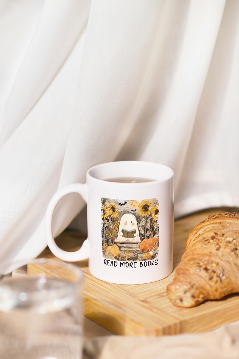 May include: White ceramic mug with a Halloween-themed illustration. The design features a cartoon ghost reading a book, surrounded by sunflowers, pumpkins, and the text "READ MORE BOOKS". The mug is on a wooden surface.