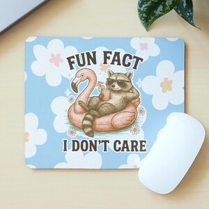 May include: A rectangular mousepad with a light blue background and white and pink flower accents. The mousepad features a raccoon wearing sunglasses, relaxing on a pink flamingo floatie, holding a drink. Text reads "FUN FACT I DON'T CARE."