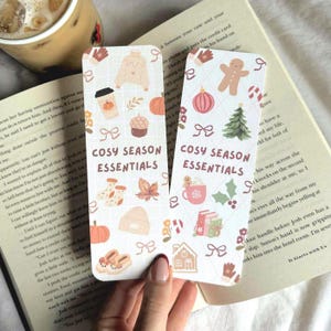 May include: Two rectangular bookmarks with a cozy season theme. One features a sweater, coffee cup, pumpkin, and other fall-themed icons. The other has a gingerbread man, Christmas tree, and other holiday icons. The text "COSY SEASON ESSENTIALS" is printed on both.