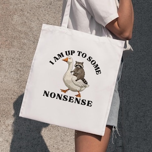 May include: White tote bag with a black graphic of a goose with a raccoon riding on its back. The text "I am up to some nonsense" is printed in black below the image.