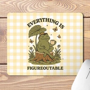 May include: A rectangular mouse mat with a yellow and white gingham pattern. The mat features a cartoon frog sitting on a tree stump, holding a cup, with the text "EVERYTHING IS FIGUREOUTABLE". Butterflies and flowers are also present.