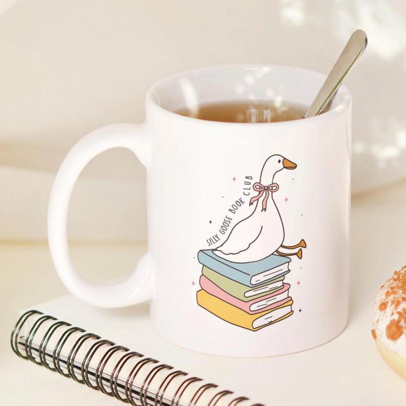 May include: White ceramic mug with a whimsical illustration of a goose wearing a pink bow, perched atop a stack of books. The mug has the text "Silly Goose Book Club". A spoon rests inside the mug, which is filled with tea.