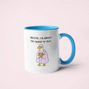 May include: White ceramic mug with a blue interior and handle. The mug features a cartoon duck wearing a pink robe and holding a golden cup. Text on the mug reads, "BESTIE, I'M ABOUT TO CRASH TF OUT."