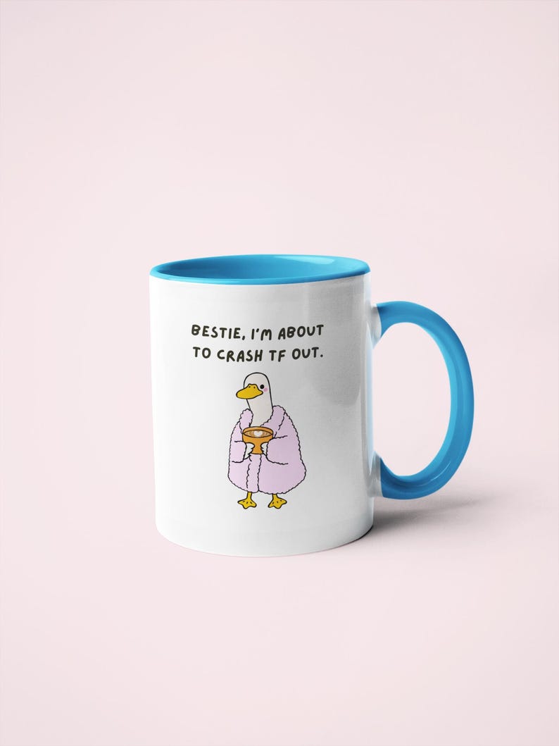 May include: White ceramic mug with a blue interior and handle. The mug features a cartoon duck wearing a pink robe and holding a golden cup. Text on the mug reads, "BESTIE, I'M ABOUT TO CRASH TF OUT."