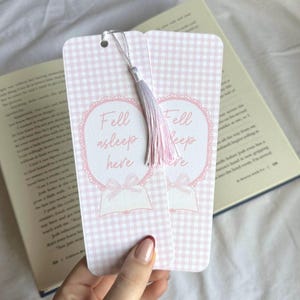 May include: Two pink and white gingham bookmarks with the text "Fell asleep here" in a heart-shaped design. Each bookmark has a silver string and a pink and lavender tassel. The bookmarks are next to an open book.