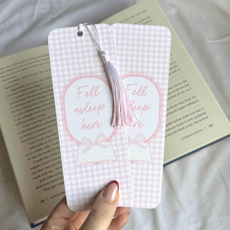 May include: Two pink and white gingham bookmarks with the text "Fell asleep here" in a heart-shaped design. Each bookmark has a silver string and a pink and lavender tassel. The bookmarks are next to an open book.