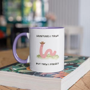 May include: White ceramic mug with a purple handle and interior. The mug features a cartoon worm wearing a cowboy hat and the text "SOMETIMES I THINK BUT THEN I FORGET". The mug is sitting on a book.