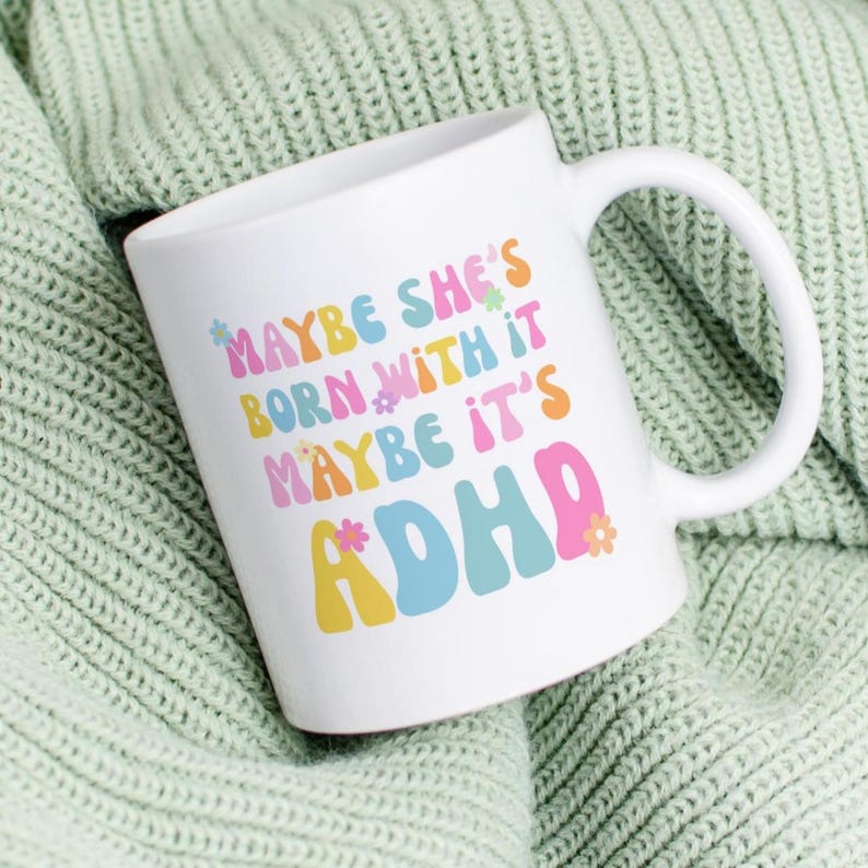 May include: White ceramic mug with the text "MAYBE SHE'S BORN WITH IT MAYBE IT'S ADHD" in colorful, retro-style lettering. The mug has a curved handle and is set against a light green knitted background.
