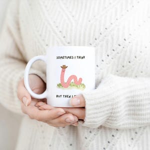 May include: White ceramic mug featuring a cartoon worm wearing a cowboy hat, with the text "Sometimes I think, but then I..." The mug is held by a person wearing a cream-colored sweater. A fun, whimsical design.