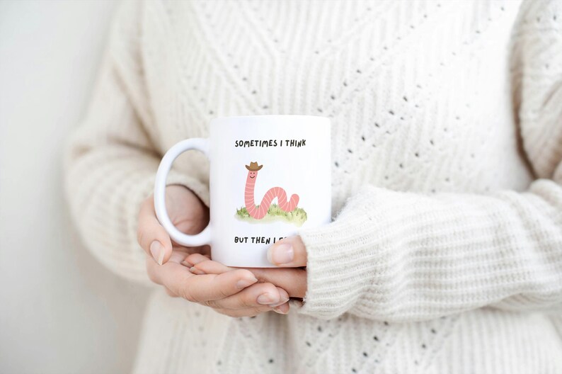 May include: White ceramic mug featuring a cartoon worm wearing a cowboy hat, with the text "Sometimes I think, but then I..." The mug is held by a person wearing a cream-colored sweater. A fun, whimsical design.