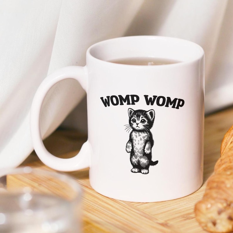 May include: White ceramic mug featuring a black and white illustration of a standing kitten. The mug has the text "WOMP WOMP" in bold, black letters above the kitten. The mug is on a wooden surface.
