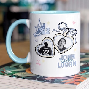 May include: A white ceramic mug with a light blue handle and interior. The exterior features a graphic of a heart-shaped locket containing two photos, a pair of ice skates with the number 22, and the text JOHN Girlie LOGAN in light blue letters.