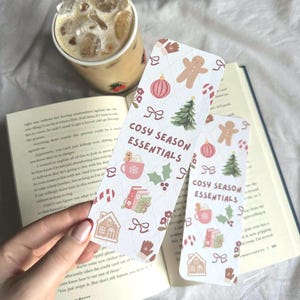 May include: Two rectangular bookmarks with a festive pattern and the text "COSY SEASON ESSENTIALS". The design includes gingerbread men, Christmas trees, and other holiday-themed illustrations. A cup of iced coffee and an open book are in the background.