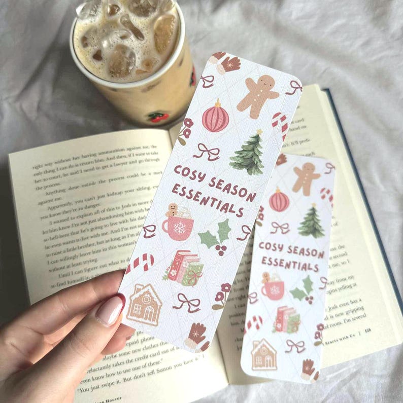 May include: Two rectangular bookmarks with a festive pattern and the text "COSY SEASON ESSENTIALS". The design includes gingerbread men, Christmas trees, and other holiday-themed illustrations. A cup of iced coffee and an open book are in the background.