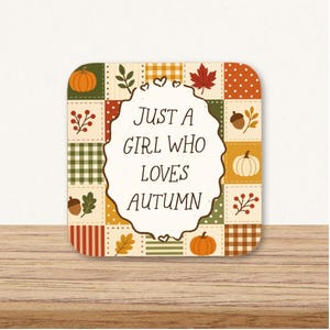 May include: A square coaster featuring a patchwork design of autumn-themed squares with pumpkins, acorns, leaves, and plaid patterns. In the centre, a scalloped white label contains the black text: JUST A GIRL WHO LOVES AUTUMN.
