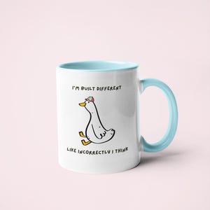 Puede incluir: Taza de cer&aacute;mica blanca con interior y asa azul claro. La taza presenta un pato de dibujos animados con sombrero, con el texto "I'M BUILT DIFFERENT LIKE INCORRECTLY I THINK."