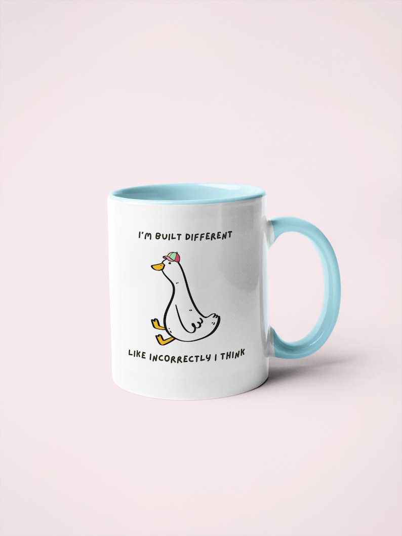 Puede incluir: Taza de cer&aacute;mica blanca con interior y asa azul claro. La taza presenta un pato de dibujos animados con sombrero, con el texto "I'M BUILT DIFFERENT LIKE INCORRECTLY I THINK."