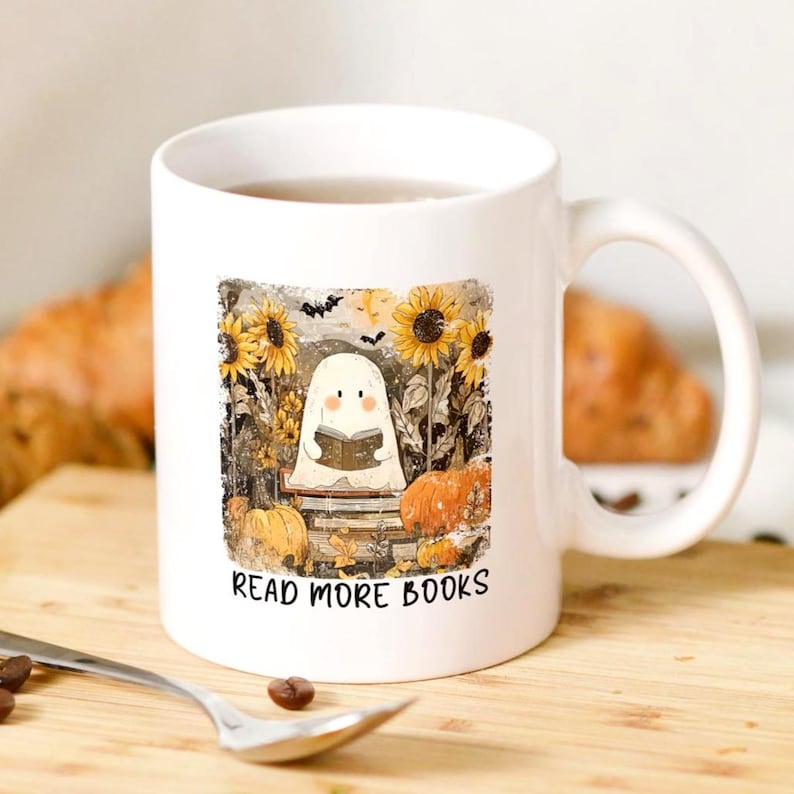 May include: White ceramic mug featuring a whimsical illustration of a ghost reading a book amidst sunflowers and pumpkins. The text "READ MORE BOOKS" is printed below the image. The mug has a curved handle.