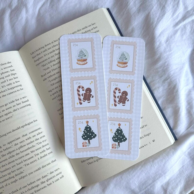 May include: Two Christmas-themed bookmarks with postage stamp designs. Each bookmark features illustrations of a snow globe, gingerbread man, candy cane, and Christmas tree. The bookmarks are set on an open book.