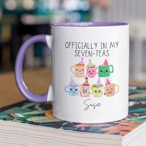May include: White ceramic mug with a purple handle and rim, featuring the text "OFFICIALLY IN MY SEVEN-TEAS" and a collection of cartoon teacups wearing party hats. The name "Susie" is written below the teacups.