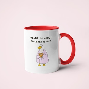 May include: White ceramic mug with a red interior and handle. The mug features a cartoon duck wearing a pink robe and holding a golden cup. Text on the mug reads: "BESTIE, I'M ABOUT TO CRASH TF OUT."