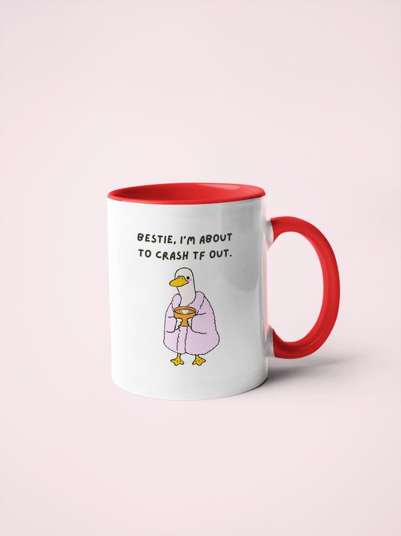 May include: White ceramic mug with a red interior and handle. The mug features a cartoon duck wearing a pink robe and holding a golden cup. Text on the mug reads: "BESTIE, I'M ABOUT TO CRASH TF OUT."