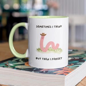 May include: White ceramic mug with a light green interior and handle. The mug features a cartoon worm wearing a cowboy hat, with the text "SOMETIMES I THINK BUT THEN I FORGET". The mug is sitting on a book.