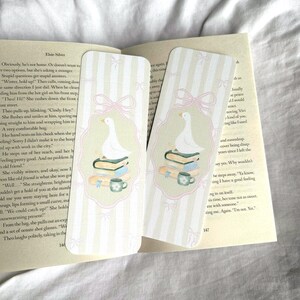 May include: Two decorative bookmarks with a whimsical design. Each bookmark features a white goose atop books and a teacup, set against a striped background with pink bows. The bookmarks are placed on an open book.