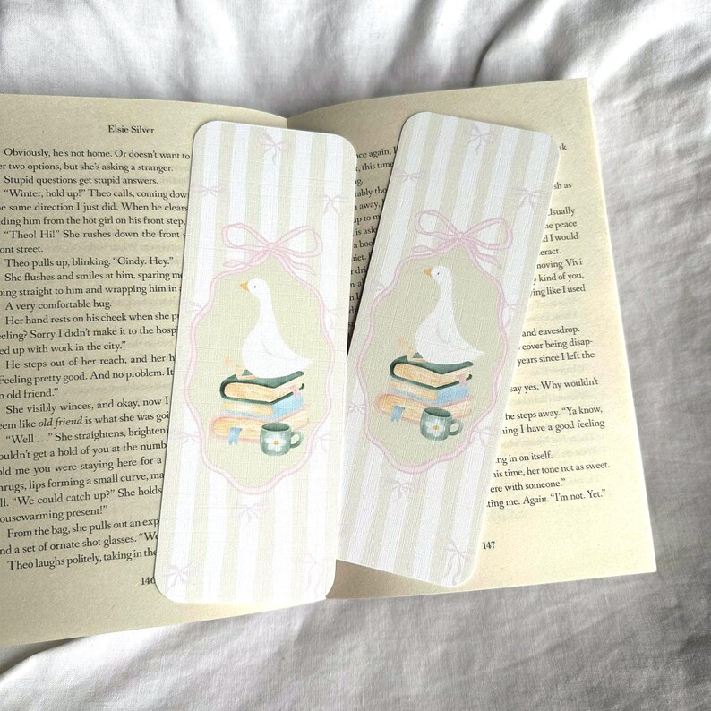 May include: Two decorative bookmarks with a whimsical design. Each bookmark features a white goose atop books and a teacup, set against a striped background with pink bows. The bookmarks are placed on an open book.